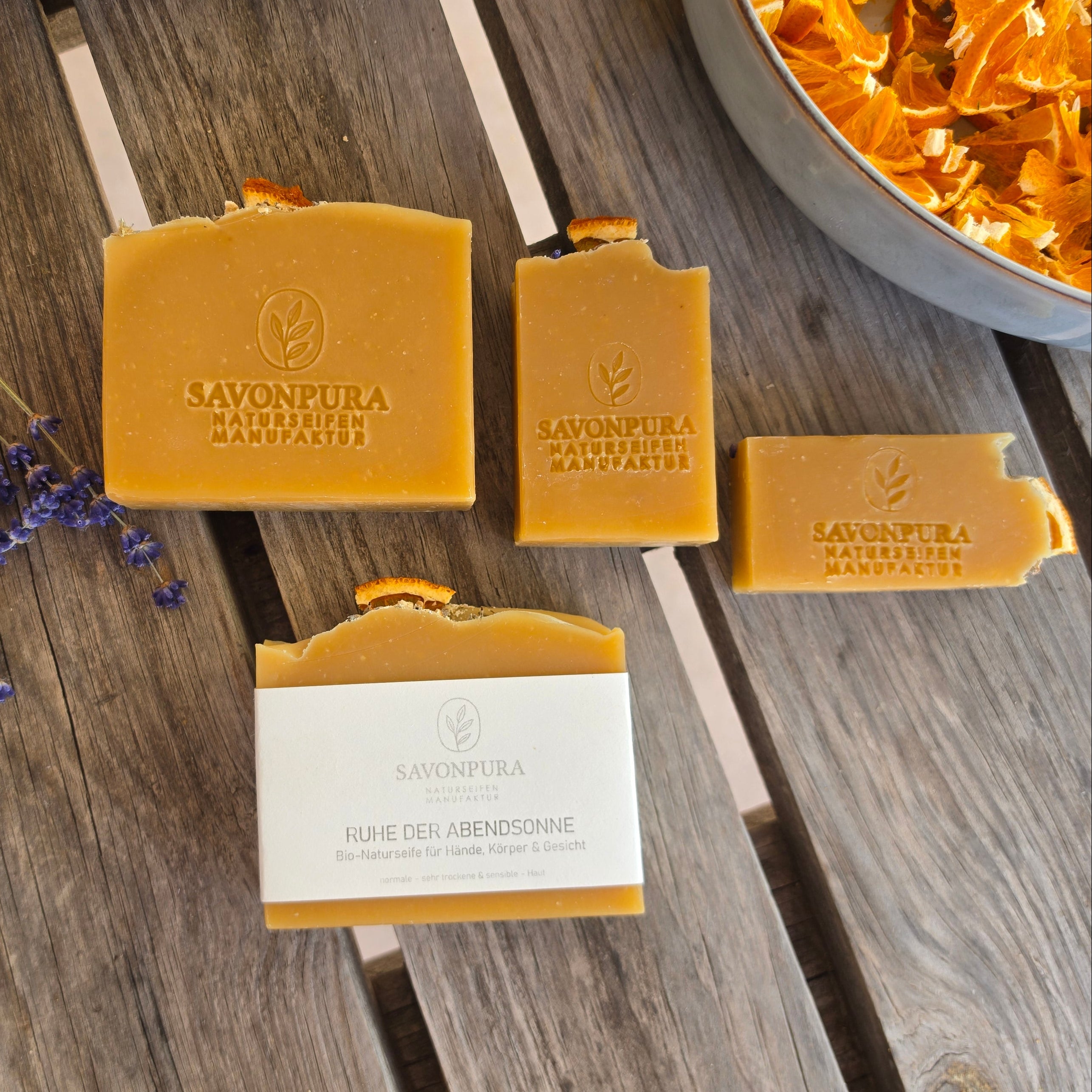 4 orangene Seifen teils mit Etikett auf Holztisch liegend mit Lavendel und Orangendeko
