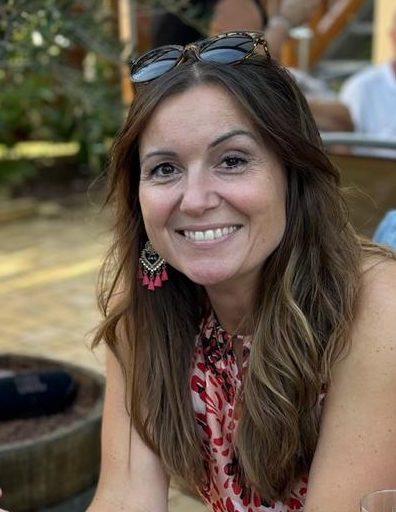 Frau mit langen, braunen Haaren und bunten Ohrringen, lächelt entspannt im Freien.